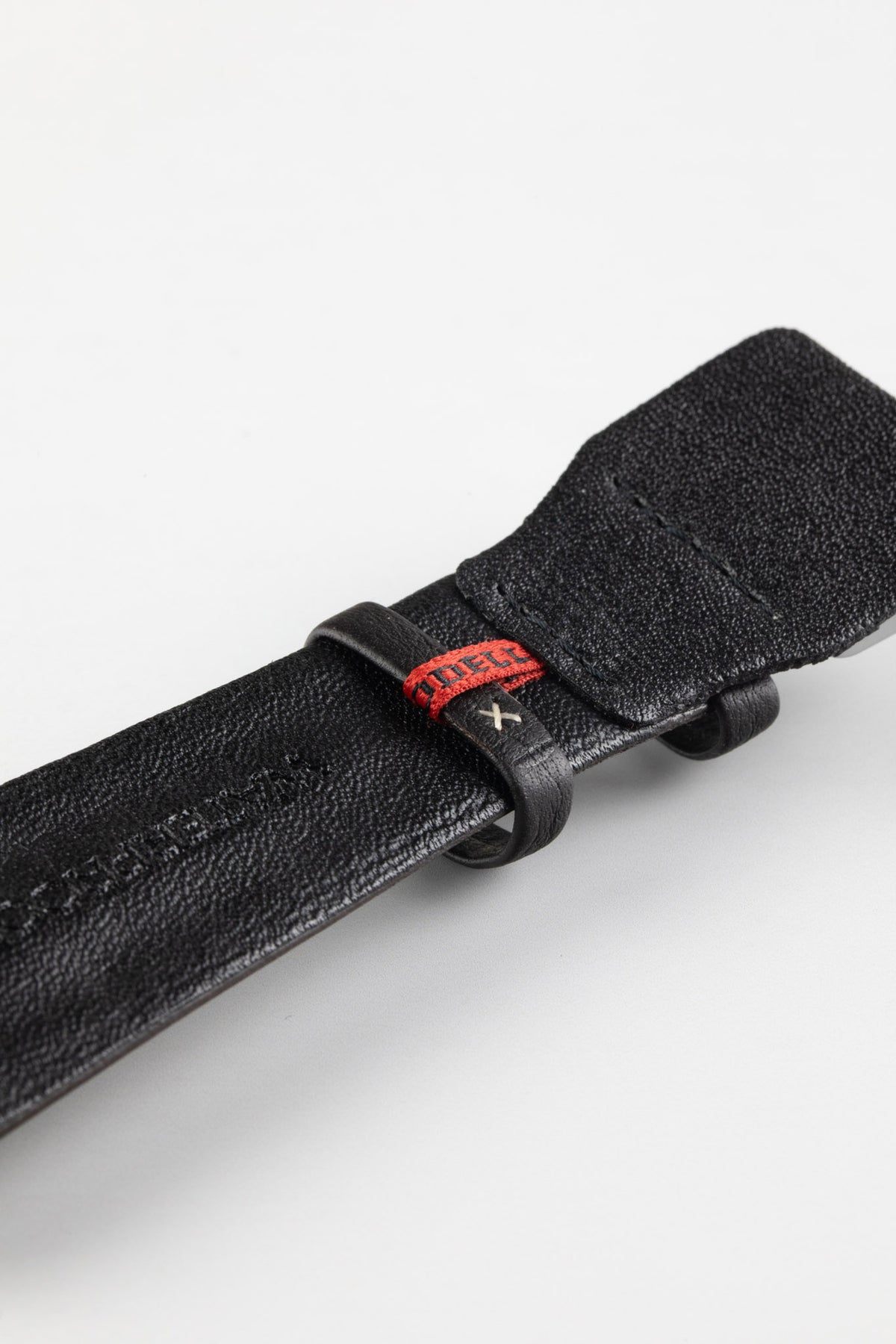 Close-up of the underside of a black padded leather rally-style watch strap, featuring a smooth black leather lining, embossed branding, dual black keeper loops, a red woven accent keeper, and reinforced lug-end padding, displayed on a light grey background to highlight the premium construction, contrasting red detailing, and motorsport-inspired design.