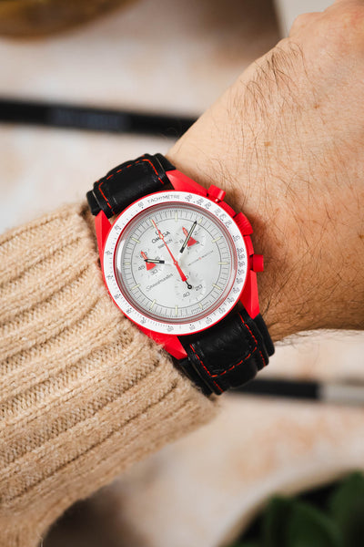 Omega x Swatch Speedmaster MoonSwatch worn on the wrist, featuring a white chronograph dial, vivid red bioceramic case, and a black padded leather rally-style watch strap with contrasting red stitching, paired with a cream knitted sweater cuff to highlight the strap's motorsport-inspired design, premium padded construction, and striking red-and-black colour combination.