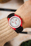 Omega x Swatch Speedmaster MoonSwatch worn on the wrist, featuring a white chronograph dial, vivid red bioceramic case, and a black padded leather rally-style watch strap with contrasting red stitching, paired with a cream knitted sweater cuff to highlight the strap's motorsport-inspired design, premium padded construction, and striking red-and-black colour combination.