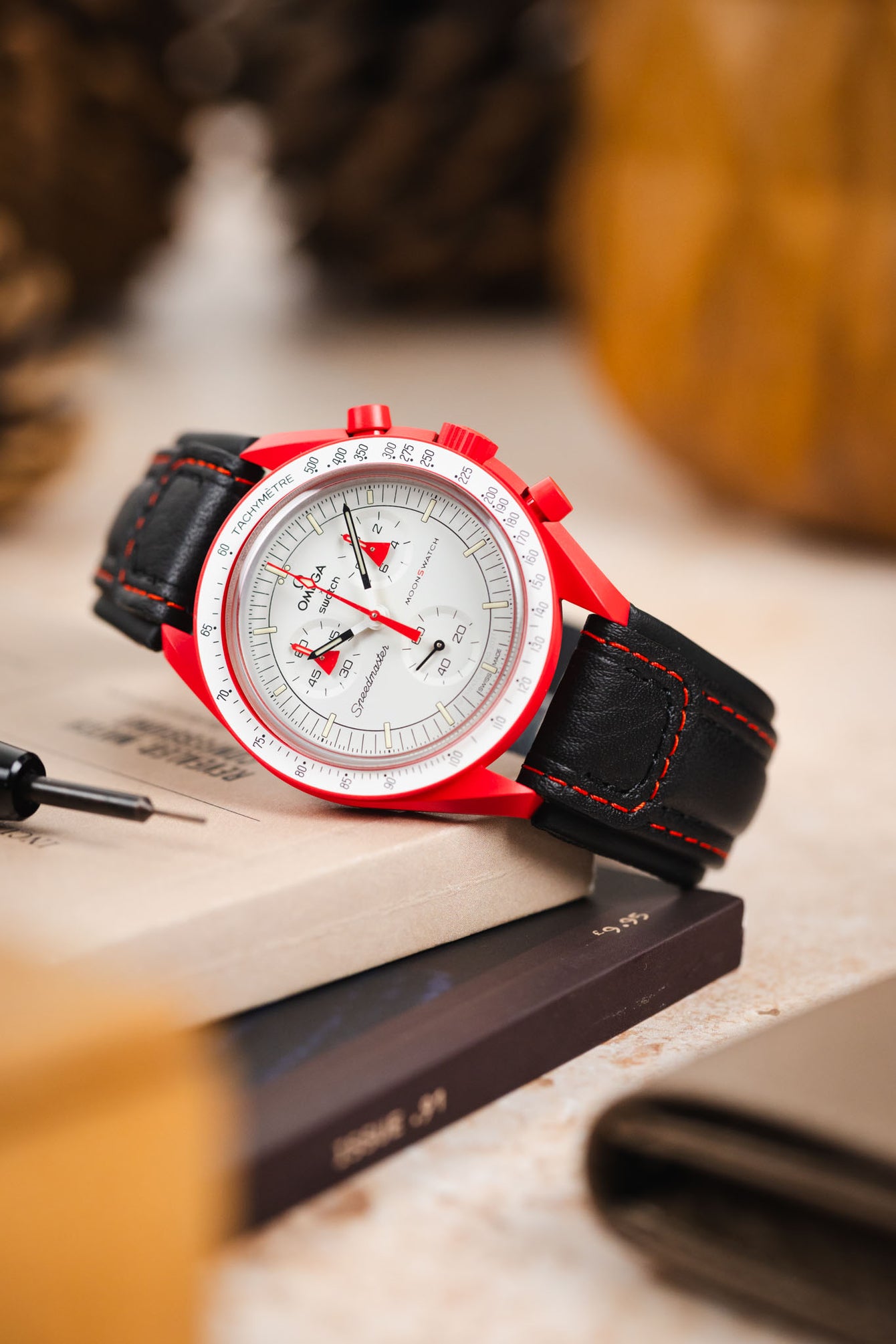 Omega x Swatch Speedmaster MoonSwatch with a white chronograph dial and vivid red bioceramic case, fitted with a black padded leather rally-style watch strap featuring contrasting red stitching, displayed on stacked books beside a spring bar tool, highlighting the strap’s motorsport-inspired design, padded construction, and bold red-and-black colour combination.