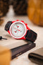 Omega x Swatch Speedmaster MoonSwatch with a white chronograph dial and vivid red bioceramic case, fitted with a black padded leather rally-style watch strap featuring contrasting red stitching, displayed on stacked books beside a spring bar tool, highlighting the strap’s motorsport-inspired design, padded construction, and bold red-and-black colour combination.