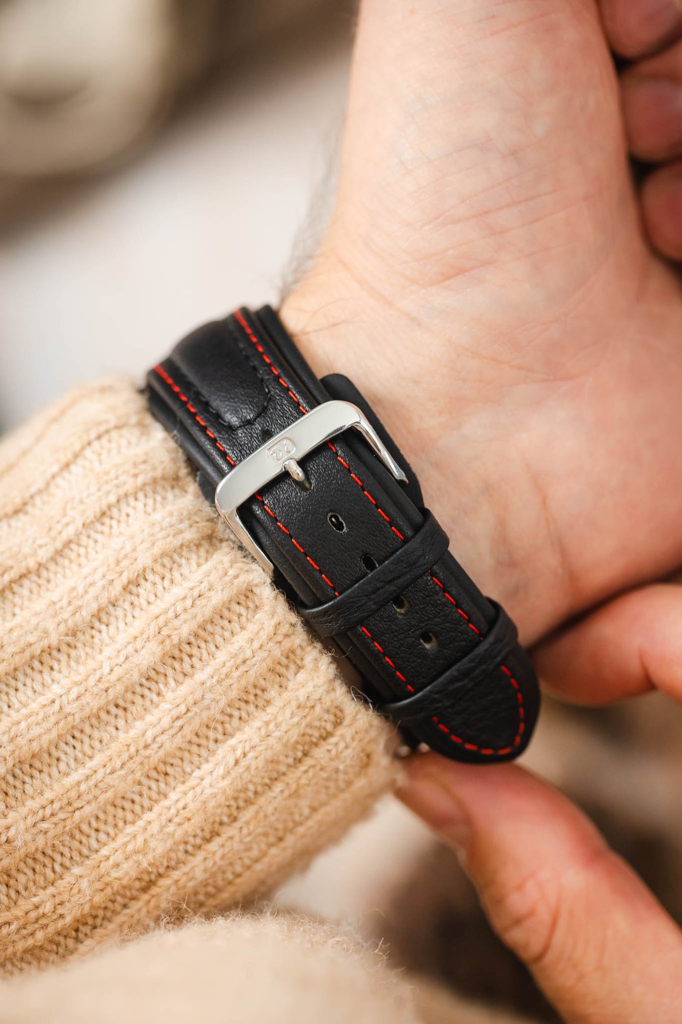 Close-up of a black padded leather watch strap worn on the wrist, featuring contrasting red stitching, dual keeper loops, integrated lug-end reinforcement panels, multiple adjustment holes, and a polished stainless steel buckle, shown with a cream knitted sweater cuff to highlight the strap's sporty motorsport-inspired design, premium leather construction, and secure fit.