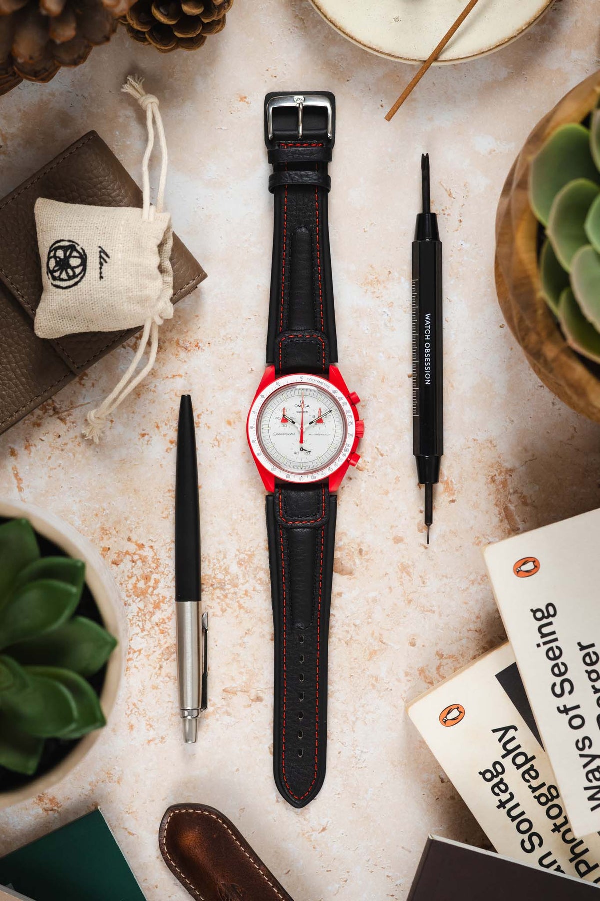 Omega x Swatch Speedmaster MoonSwatch with a white dial, red bioceramic case, and black padded leather rally-style watch strap featuring contrasting red stitching, displayed on a stone surface alongside a Watch Obsession spring bar tool, pen, books, leather accessories, and succulent plants, highlighting the sporty motorsport-inspired strap design and bold red-and-black colour combination.