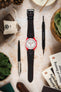 Omega x Swatch Speedmaster MoonSwatch with a white dial, red bioceramic case, and black padded leather rally-style watch strap featuring contrasting red stitching, displayed on a stone surface alongside a Watch Obsession spring bar tool, pen, books, leather accessories, and succulent plants, highlighting the sporty motorsport-inspired strap design and bold red-and-black colour combination.