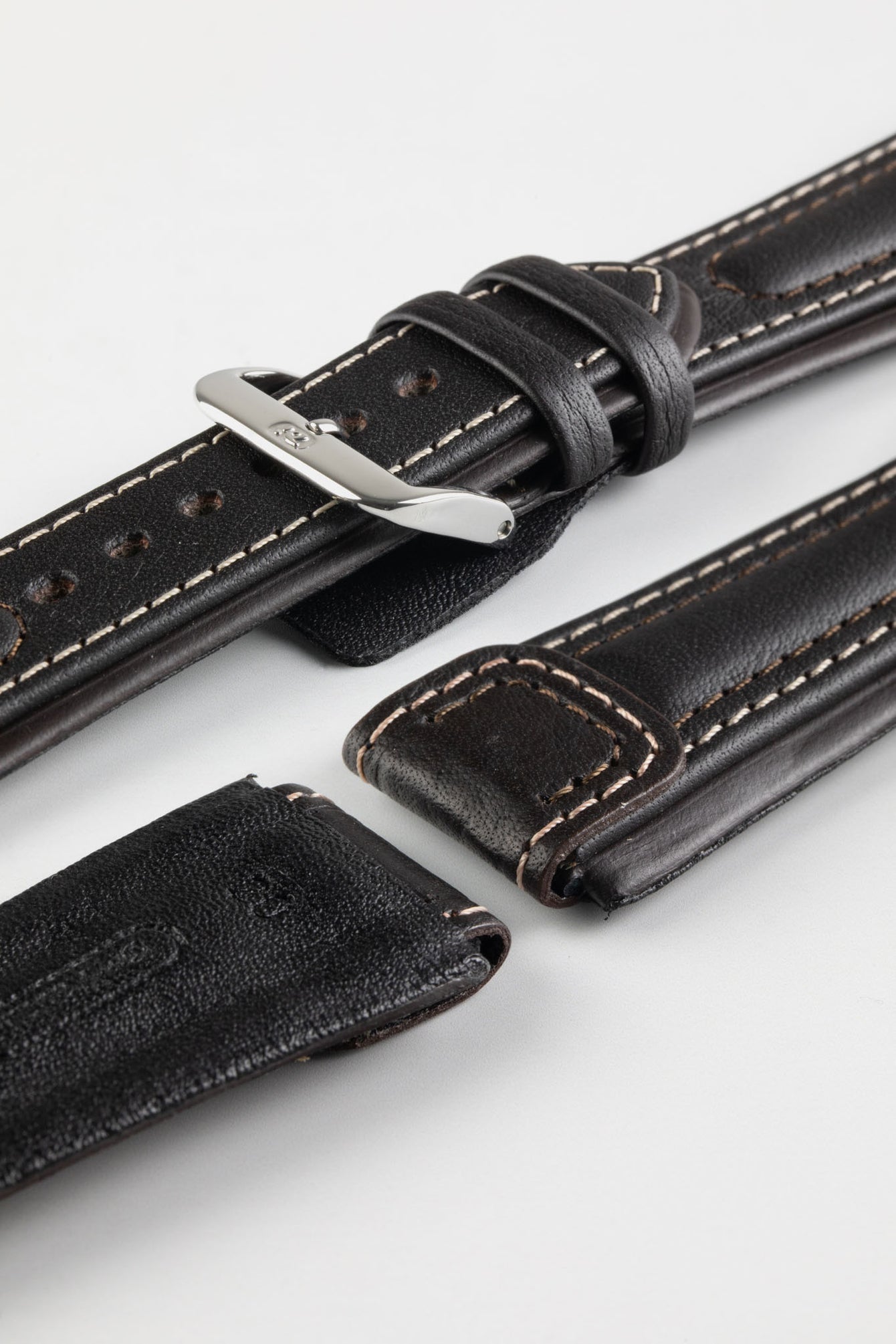 Close-up detail view of a dark brown padded leather rally-style watch strap featuring cream contrast stitching, a polished stainless steel buckle, dual keeper loops, integrated lug-end reinforcement panels, and a black leather lining with embossed branding, displayed on a light grey background with one strap section detached to highlight the premium handcrafted construction, cushioned profile, and vintage motorsport-inspired design.