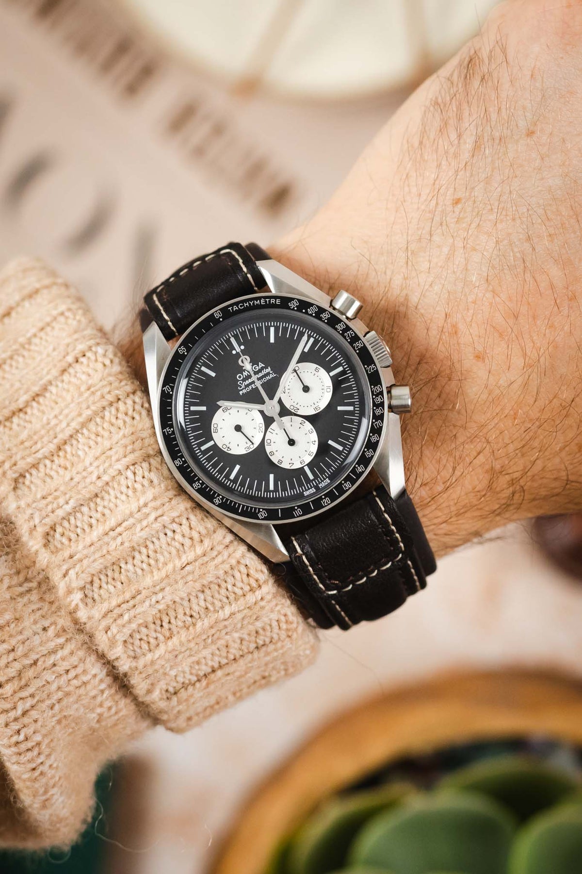 Omega Speedmaster Professional chronograph watch worn on the wrist, featuring a black dial with three white chronograph sub-dials, black tachymeter bezel, and a dark brown padded leather rally-style watch strap with cream contrast stitching and integrated lug-end reinforcement panels, paired with a cream knitted sweater cuff to highlight the strap's premium handcrafted construction, cushioned profile, and vintage motorsport-inspired design.