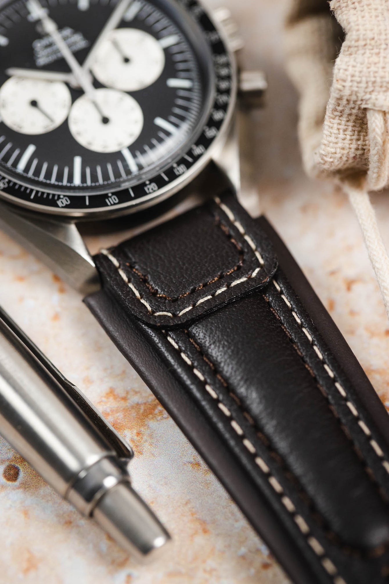 Close-up detail of an Omega Speedmaster Professional chronograph fitted with a dark brown padded leather rally-style watch strap, featuring cream contrast stitching and integrated lug-end reinforcement panels, displayed alongside a metal pen on a stone-effect surface to highlight the strap’s supple leather texture, cushioned construction, and vintage motorsport-inspired design.