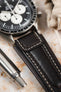 Close-up detail of an Omega Speedmaster Professional chronograph fitted with a dark brown padded leather rally-style watch strap, featuring cream contrast stitching and integrated lug-end reinforcement panels, displayed alongside a metal pen on a stone-effect surface to highlight the strap’s supple leather texture, cushioned construction, and vintage motorsport-inspired design.