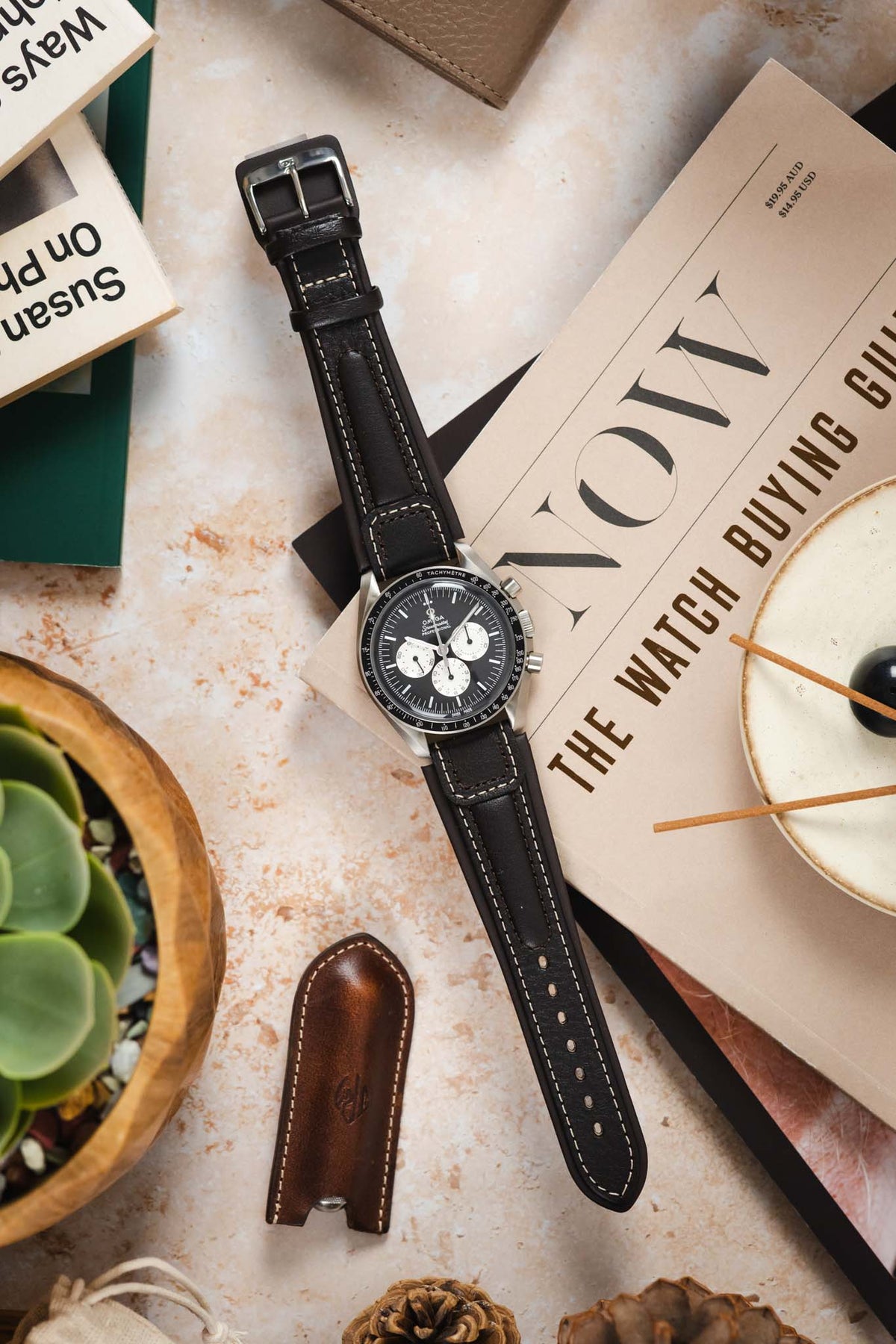 Omega Speedmaster Professional chronograph watch fitted with a dark brown padded leather rally-style watch strap featuring cream contrast stitching, displayed across a NOW: The Watch Buying Guide magazine alongside a leather watch pouch, succulent plant, and decorative accessories, highlighting the strap's vintage motorsport-inspired design, cushioned construction, and refined handcrafted leather finish.
