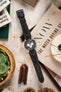 Omega Speedmaster Professional chronograph watch fitted with a dark brown padded leather rally-style watch strap featuring cream contrast stitching, displayed across a NOW: The Watch Buying Guide magazine alongside a leather watch pouch, succulent plant, and decorative accessories, highlighting the strap's vintage motorsport-inspired design, cushioned construction, and refined handcrafted leather finish.
