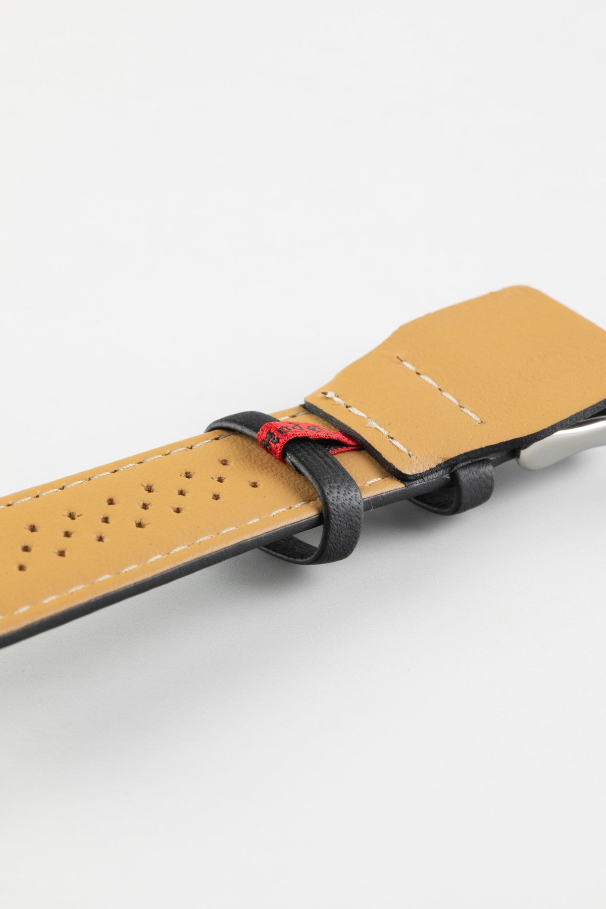 Black leather watch strap with a beige perforated leather lining, contrast stitching, and a red branded tab, shown in close-up on a white background.