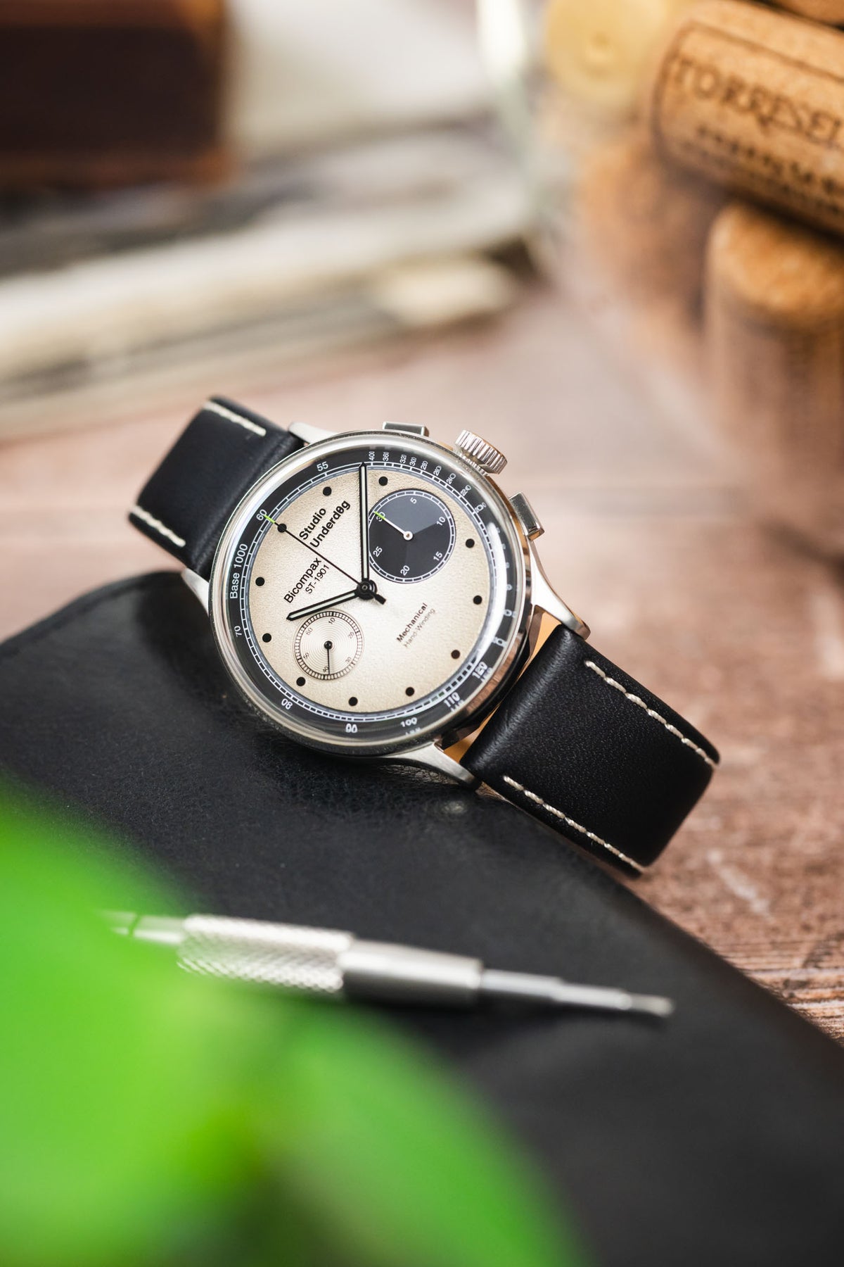 Studio Underd0g Bicompax chronograph watch on a black leather strap with beige contrast stitching, displayed on a leather notebook with watchmaking tools and vintage accessories.