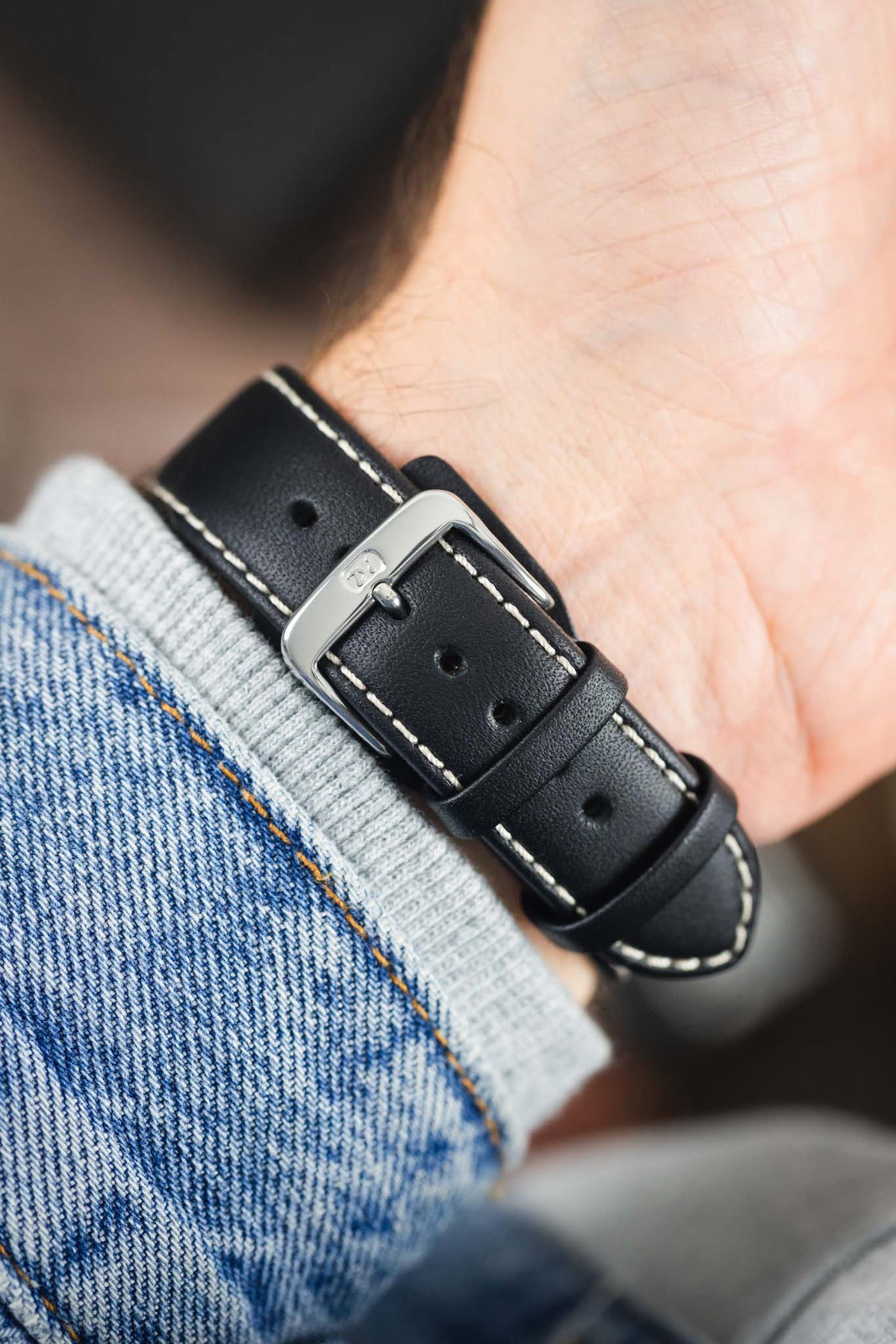 Black leather watch strap with beige contrast stitching and a polished stainless steel buckle, shown on wrist from the underside.