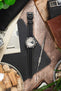Studio Underd0g Bicompax chronograph watch on a black leather strap with beige contrast stitching, displayed with vintage accessories on a rustic wooden surface.