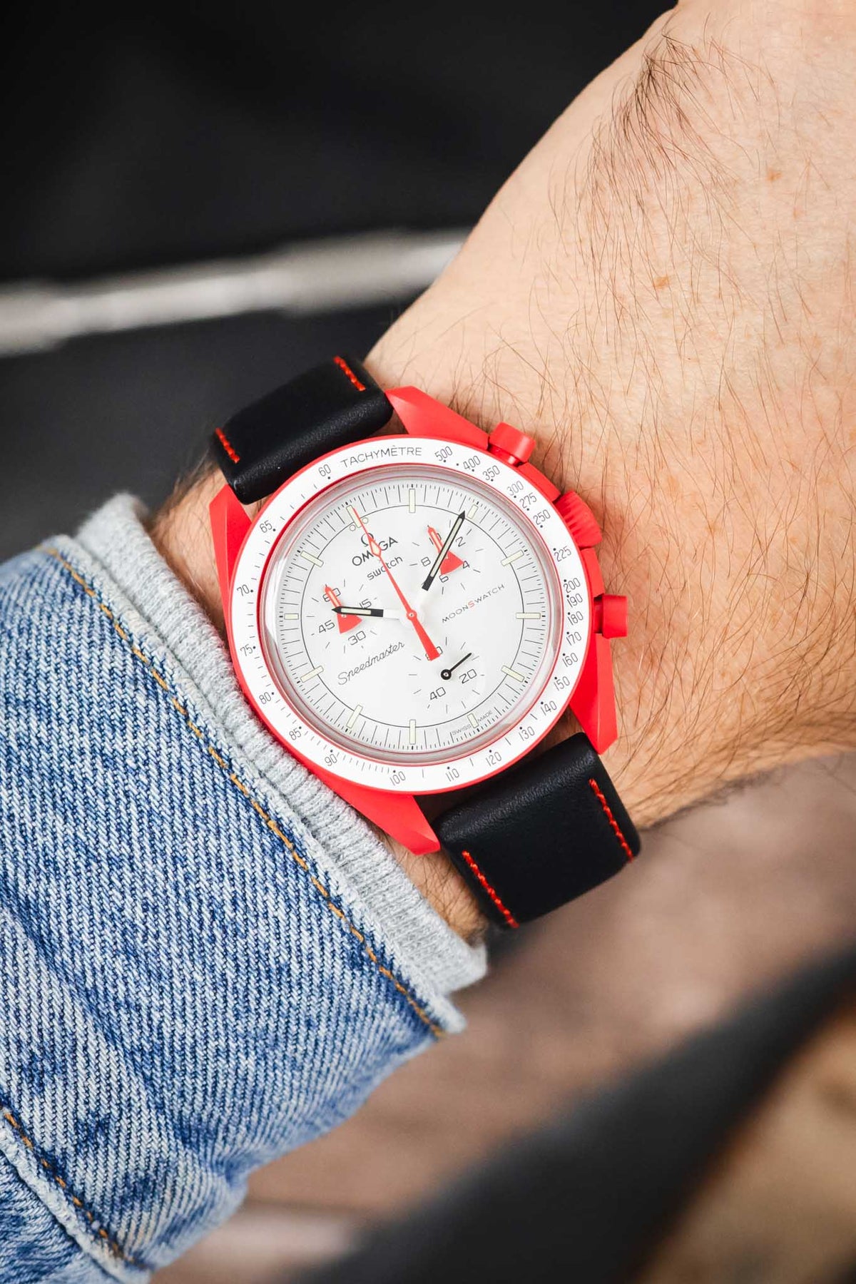 Omega x Swatch MoonSwatch chronograph on a black leather strap with red contrast stitching, shown on wrist with a red case and white dial.