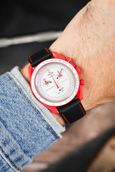 Omega x Swatch MoonSwatch chronograph on a black leather strap with red contrast stitching, shown on wrist with a red case and white dial.