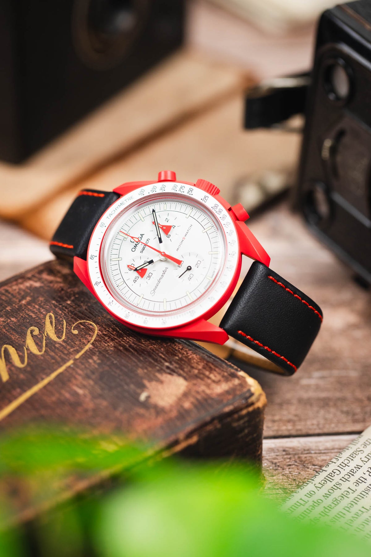 Omega x Swatch MoonSwatch chronograph on a black leather strap with red contrast stitching, displayed on a vintage wooden box with retro camera accessories.