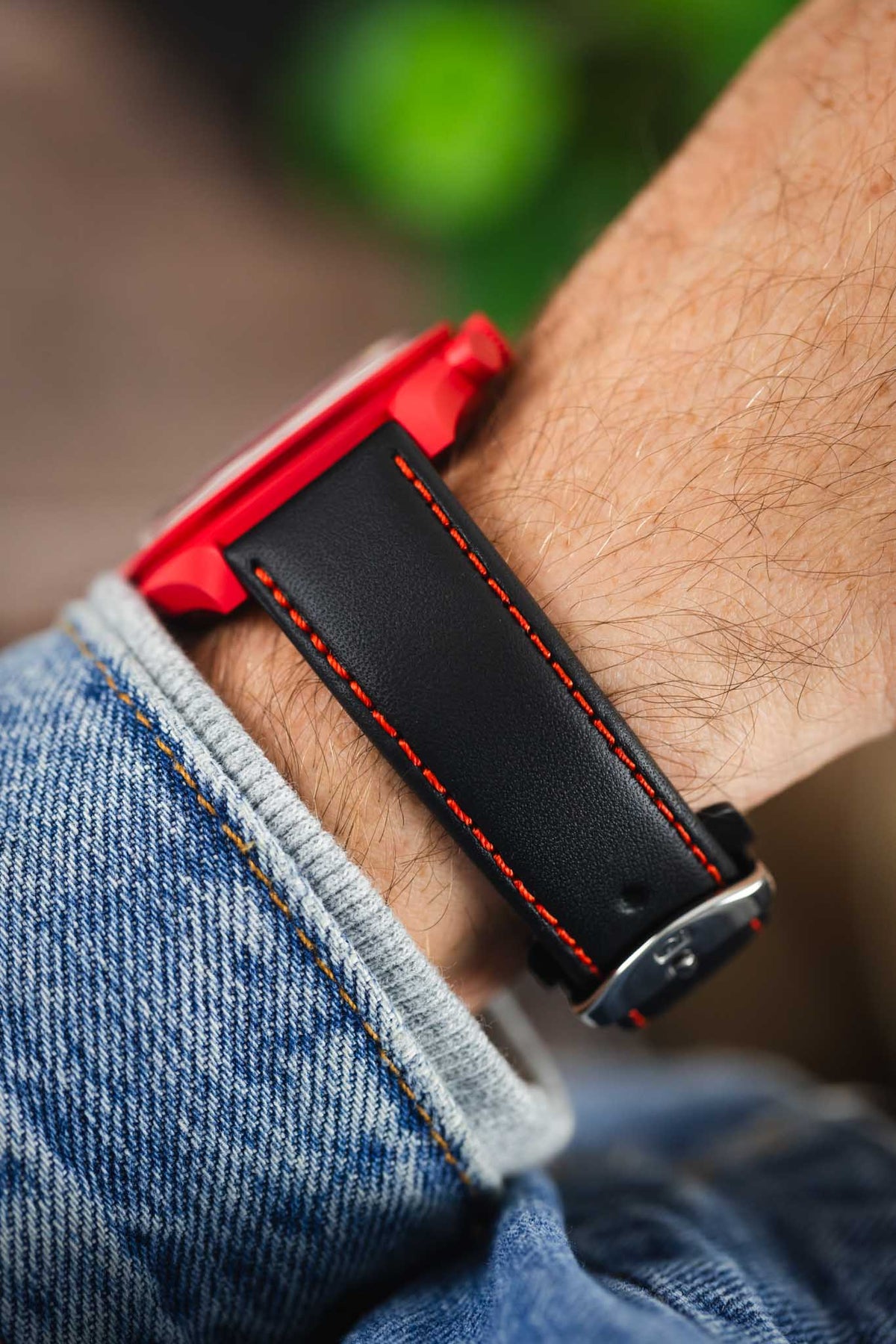 Black leather watch strap with red contrast stitching and a polished stainless steel buckle, shown on wrist with a red Omega x Swatch MoonSwatch chronograph.