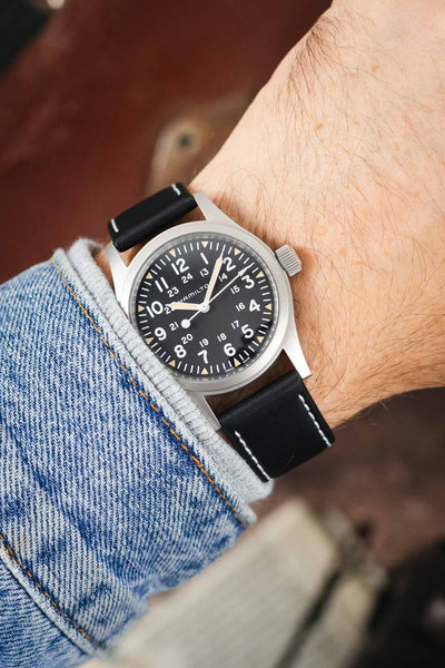 Hamilton Khaki Field-style watch on a black leather strap with white contrast stitching, shown on wrist with a black dial and brushed stainless steel case.