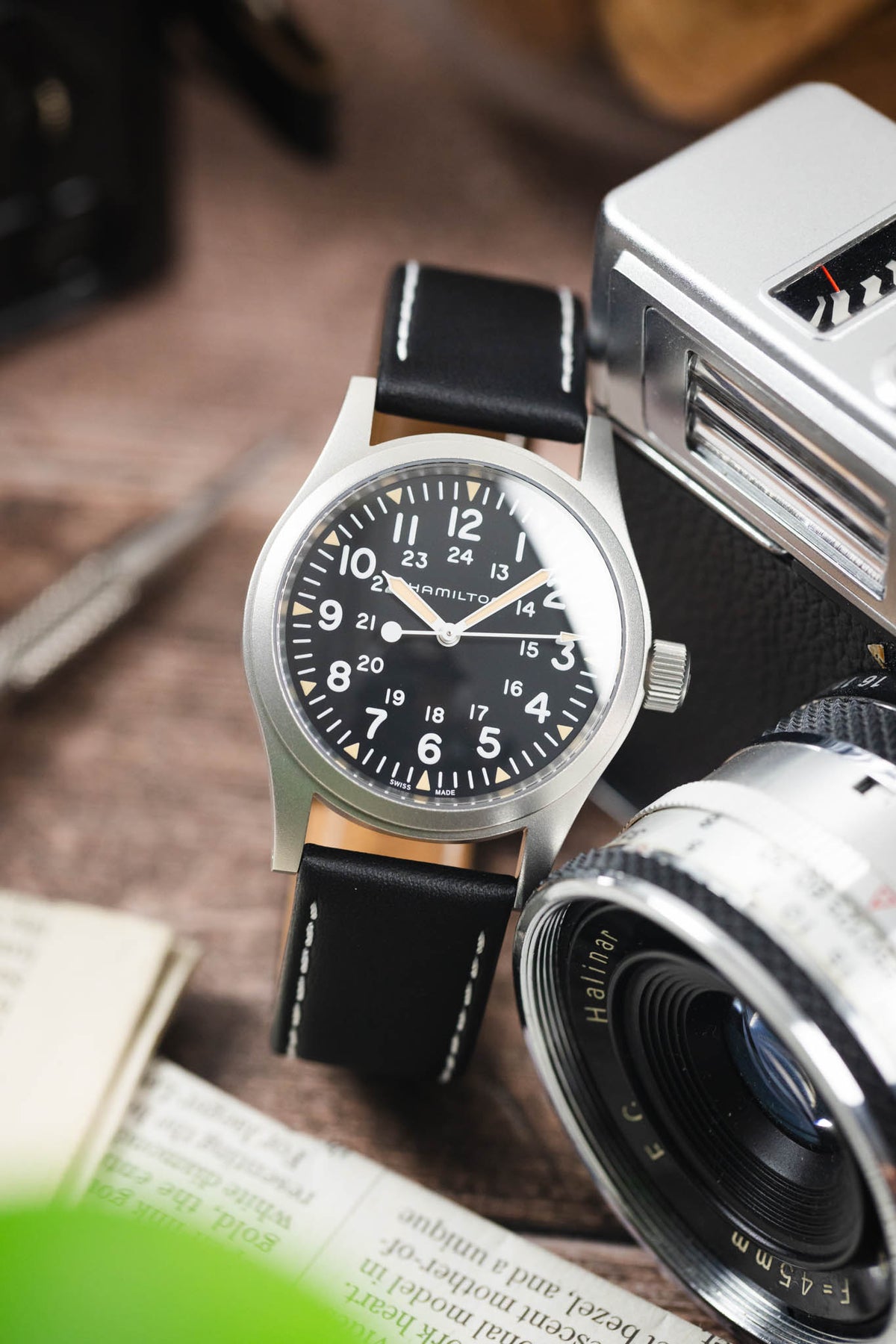 Hamilton Khaki Field-style watch on a black leather strap with white contrast stitching, displayed beside a vintage camera and lens on a rustic wooden surface.