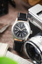 Hamilton Khaki Field-style watch on a black leather strap with white contrast stitching, displayed beside a vintage camera and lens on a rustic wooden surface.