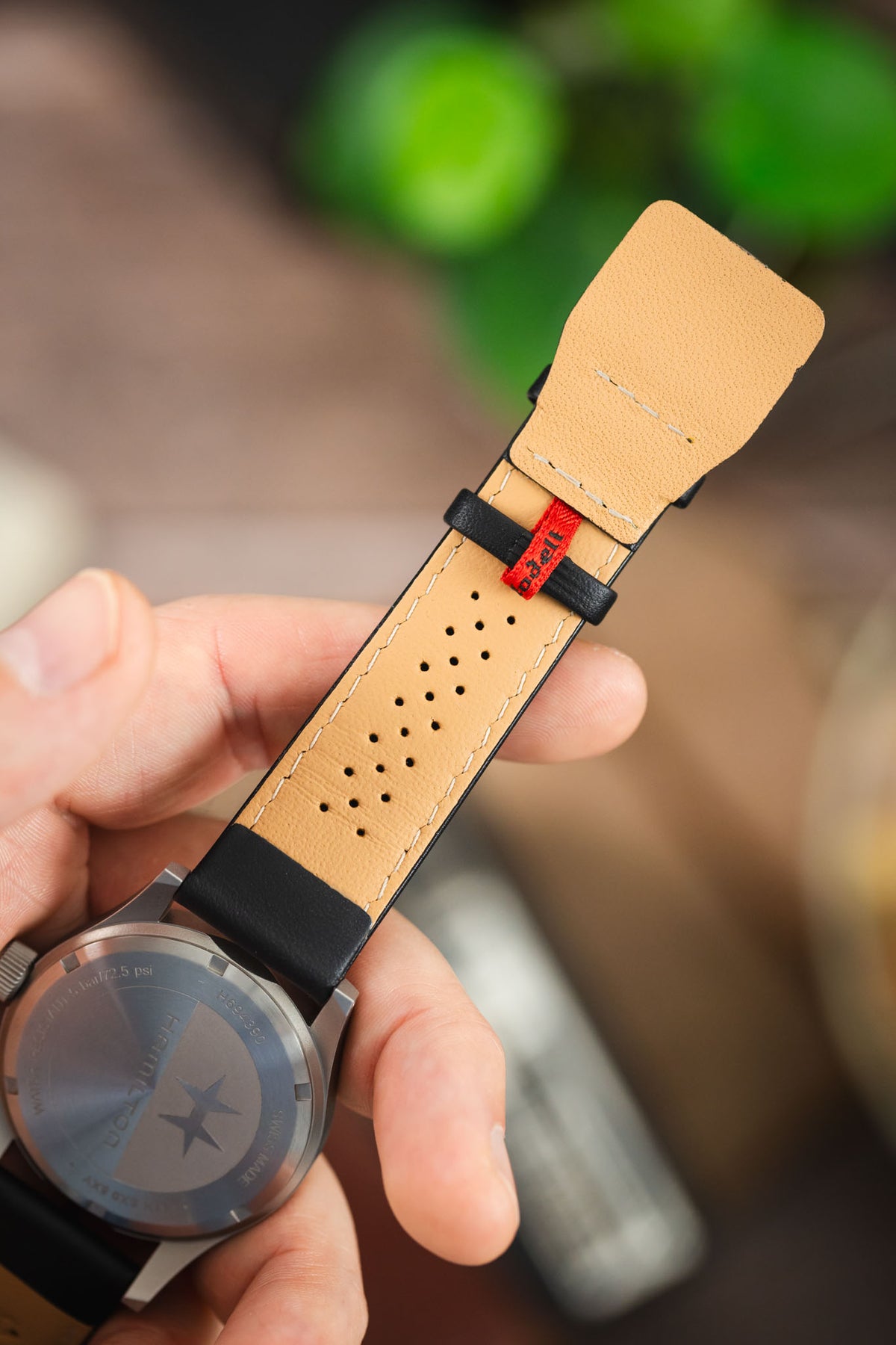Black leather watch strap with a beige perforated leather lining, white contrast stitching, and a red branded tab, shown fitted to a Hamilton field watch.
