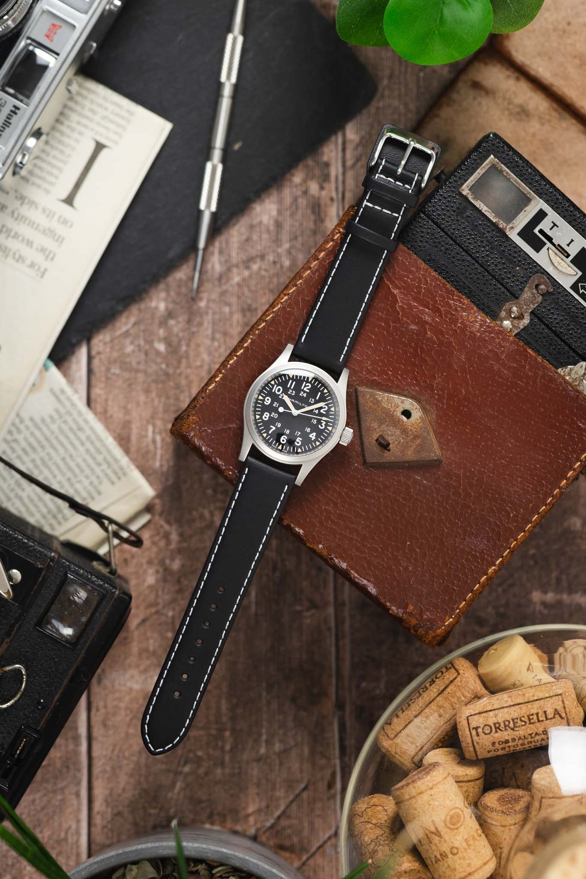 Hamilton Khaki Field-style watch on a black leather strap with white contrast stitching, displayed with vintage accessories on a rustic wooden surface.