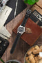 Hamilton Khaki Field-style watch on a black leather strap with white contrast stitching, displayed with vintage accessories on a rustic wooden surface.