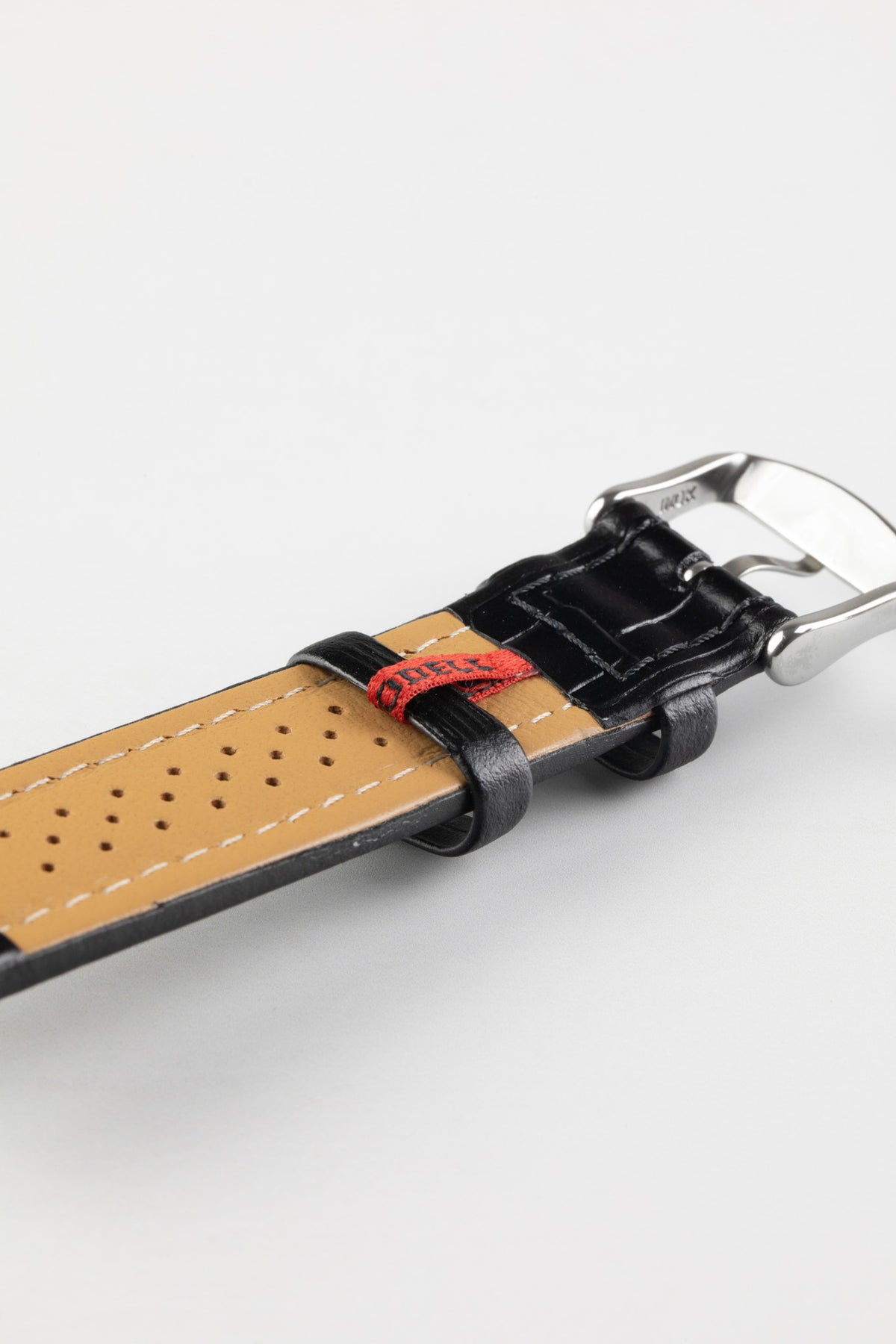 Black alligator-grain leather watch strap with a perforated beige leather lining, white stitching, and a polished stainless steel buckle, shown in close-up on a white background.