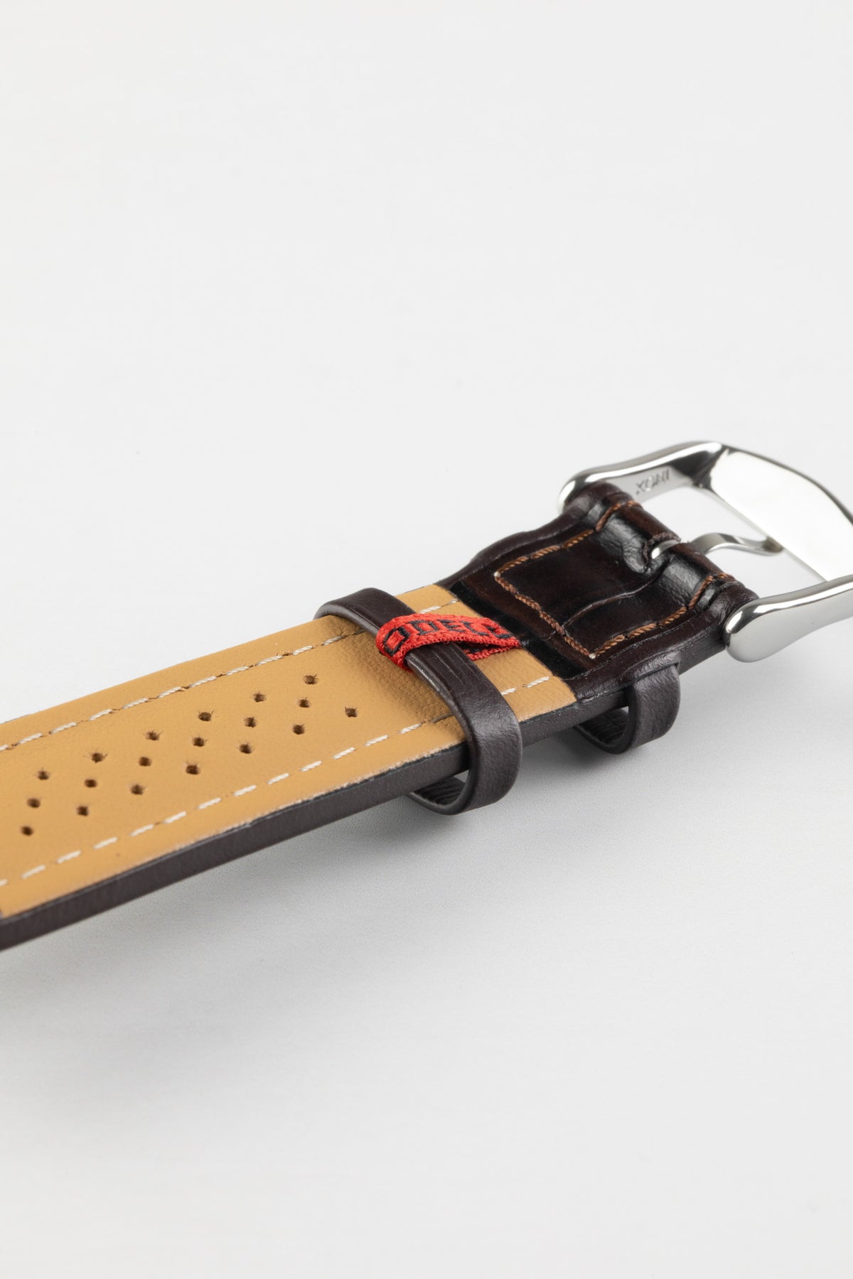 Dark brown alligator-grain leather watch strap with a perforated beige leather lining, white stitching, and a polished stainless steel buckle, shown in close-up on a white background.