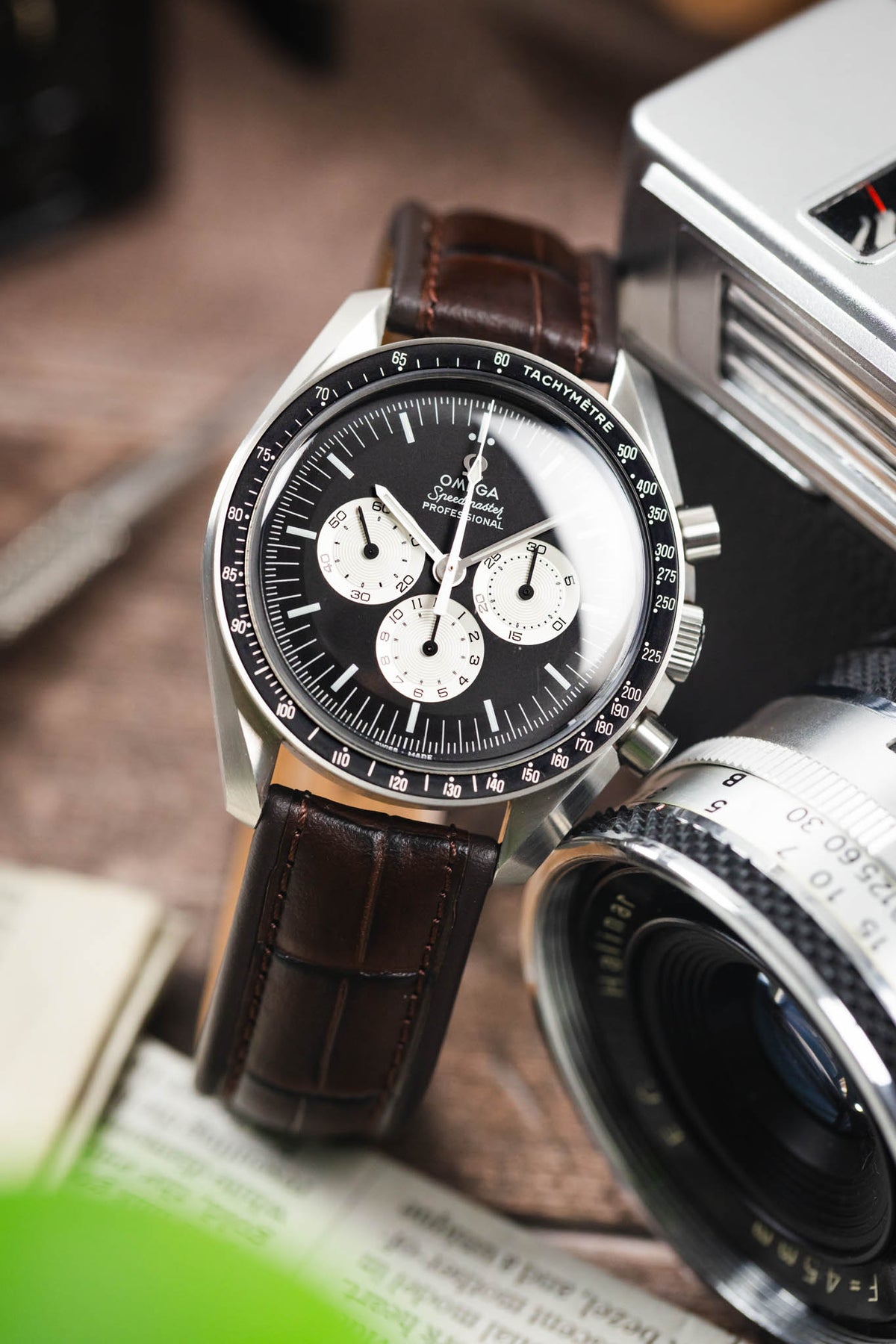 Omega Speedmaster Professional chronograph on a dark brown alligator-grain leather strap, displayed beside a vintage camera and lens on a rustic wooden surface.