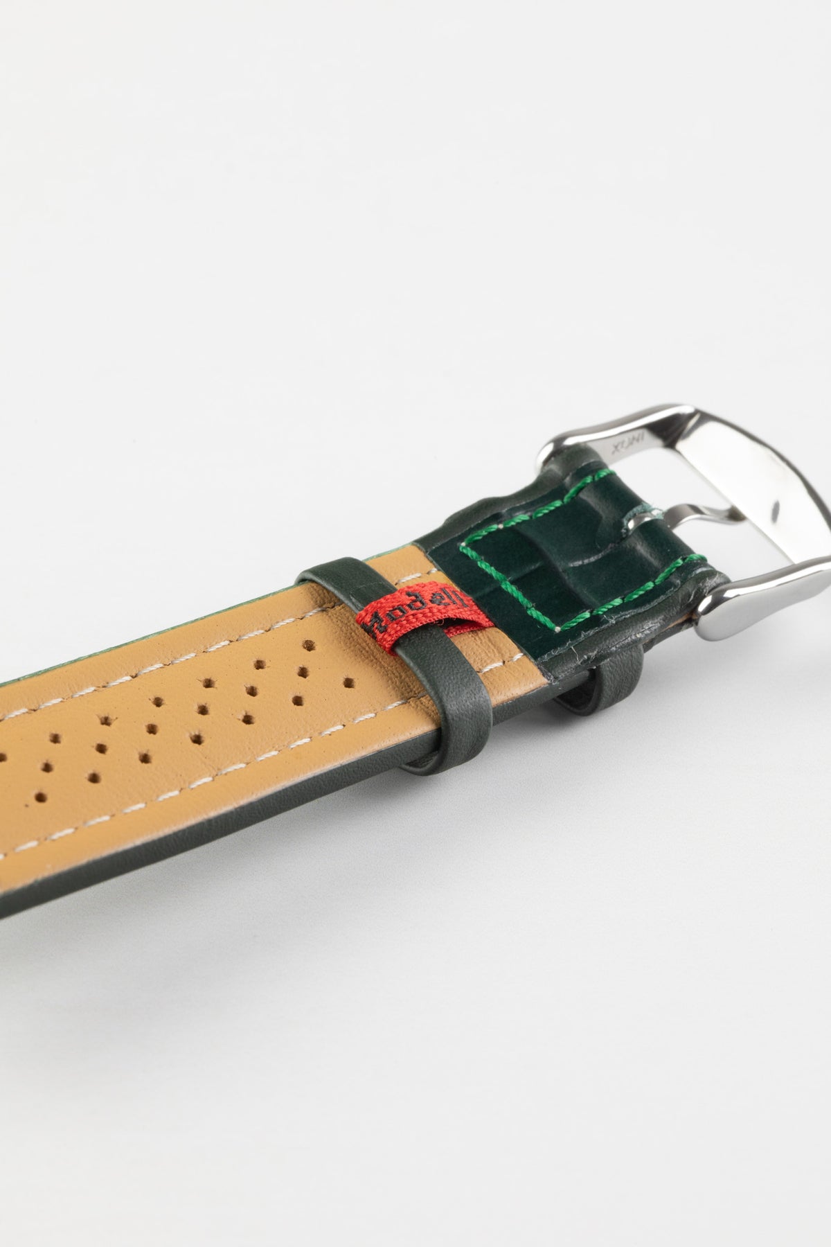 Close-up product photograph of a dark green alligator-grain leather watch strap displayed on a white background. The image highlights the underside of the strap, featuring a beige perforated leather lining, contrasting white stitching, dark green leather keepers, a polished stainless steel pin buckle, and a red branded fabric tab. The embossed crocodile-style grain and padded construction showcase the strap’s premium craftsmanship, elegant styling, and comfortable wear.