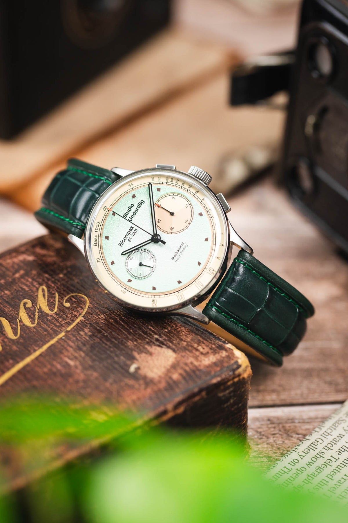 Studio Underd0g Bicompax chronograph watch on a dark green alligator-grain leather strap, displayed on a vintage wooden box with retro camera accessories.