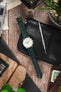 Studio Underd0g Bicompax chronograph watch on a dark green alligator-grain leather strap, displayed with vintage accessories and watchmaking tools on a rustic wooden surface.