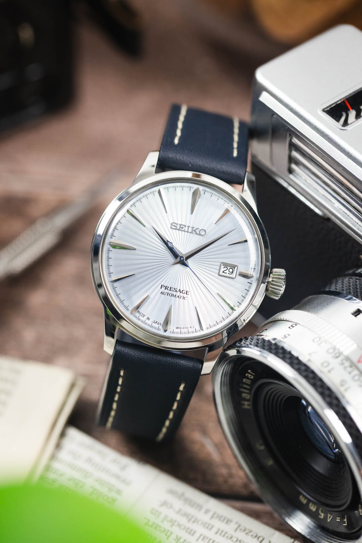 Seiko Presage automatic watch on a navy blue leather strap with cream contrast stitching, displayed beside a vintage camera and lens on a rustic wooden surface.