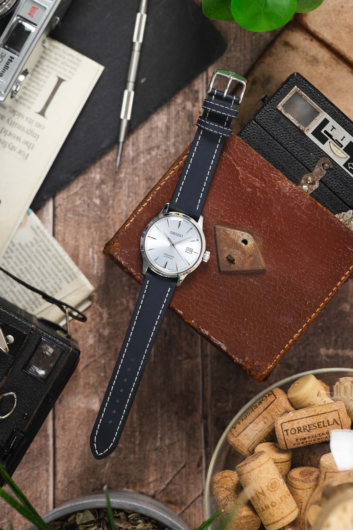 Seiko Presage automatic watch on a navy blue leather strap with cream contrast stitching, displayed with vintage accessories on a rustic wooden surface.