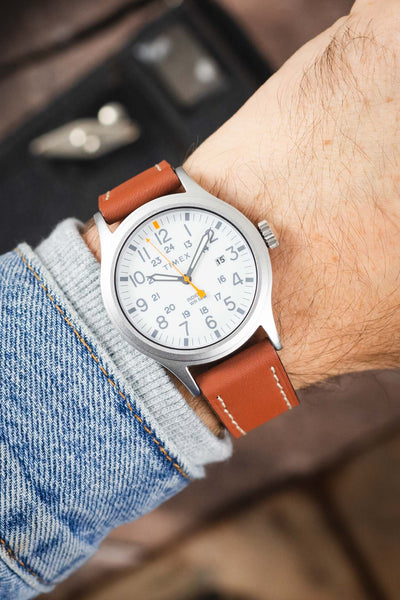 Timex Expedition-style field watch on a tan leather strap with cream contrast stitching, shown on wrist with a white dial and brushed grey case.
