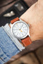 Timex Expedition-style field watch on a tan leather strap with cream contrast stitching, shown on wrist with a white dial and brushed grey case.