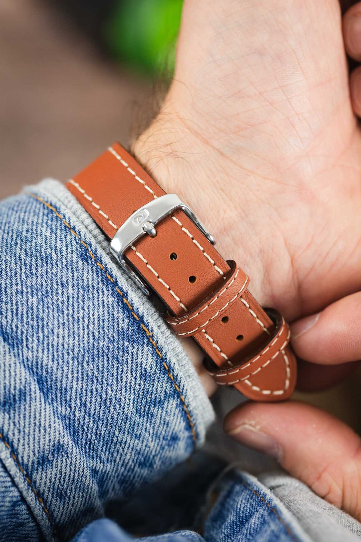 Tan leather watch strap with cream contrast stitching and a polished stainless steel buckle, shown on wrist from the underside.