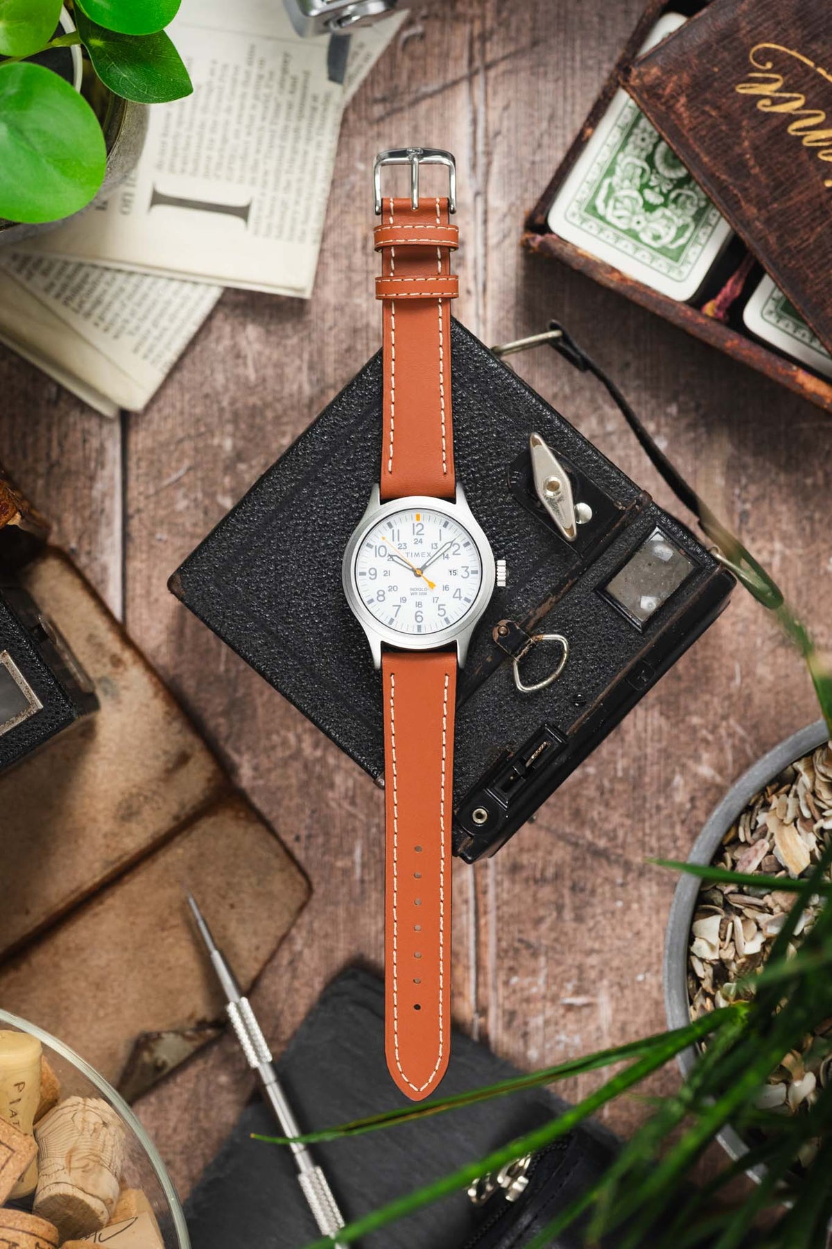 Timex Expedition-style field watch on a tan leather strap with cream contrast stitching, displayed with vintage accessories on a rustic wooden surface.