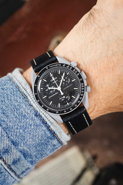 Omega x Swatch MoonSwatch chronograph on a black leather strap with cream contrast stitching, shown on wrist with a black dial and grey case.