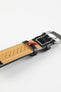 Close-up product photograph of a black vintage-style leather rally watch strap displayed on a white background. The image highlights the underside of the strap, featuring a beige perforated leather lining, contrasting cream stitching, black leather keepers, a polished stainless steel pin buckle, and a red branded fabric tab. The distressed leather finish and perforated rally design showcase the strap's motorsport-inspired styling, breathable construction, and handcrafted character.