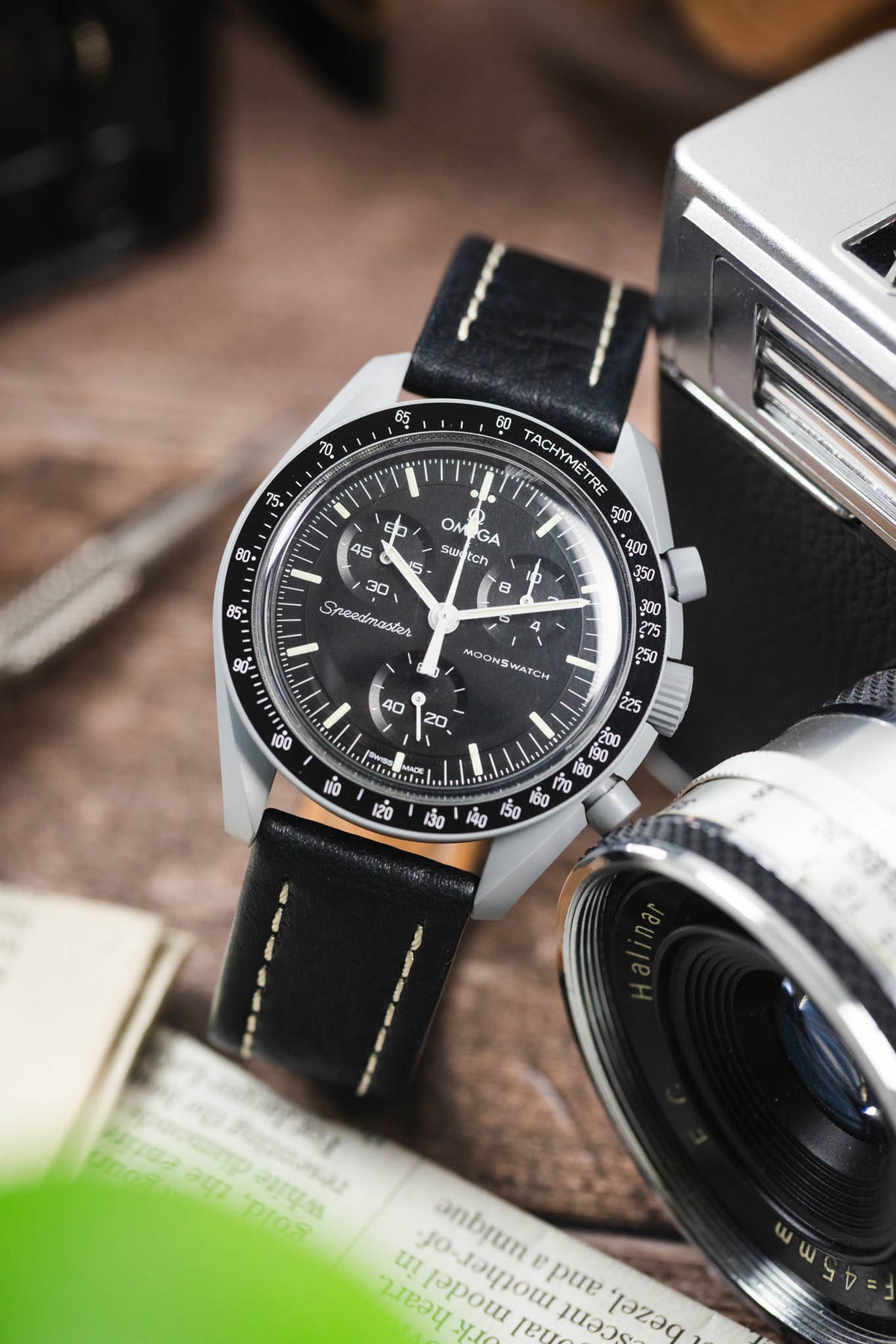 Omega x Swatch MoonSwatch chronograph on a black leather strap with cream contrast stitching, displayed beside a vintage camera and lens on a rustic wooden surface.