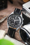 Omega x Swatch MoonSwatch chronograph on a black leather strap with cream contrast stitching, displayed beside a vintage camera and lens on a rustic wooden surface.
