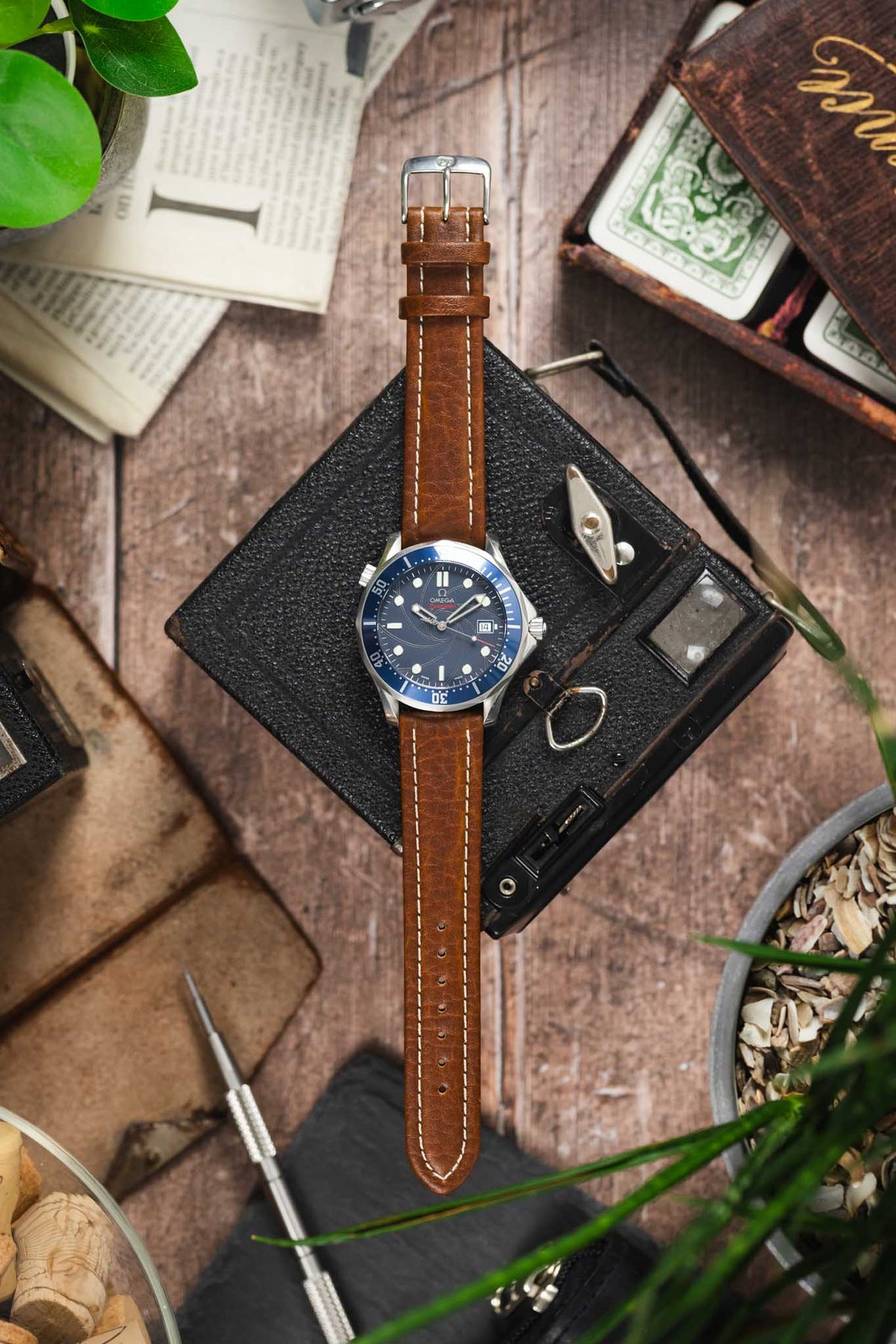 Omega Seamaster Professional dive watch on a brown leather strap with cream contrast stitching, displayed with vintage accessories on a rustic wooden surface.