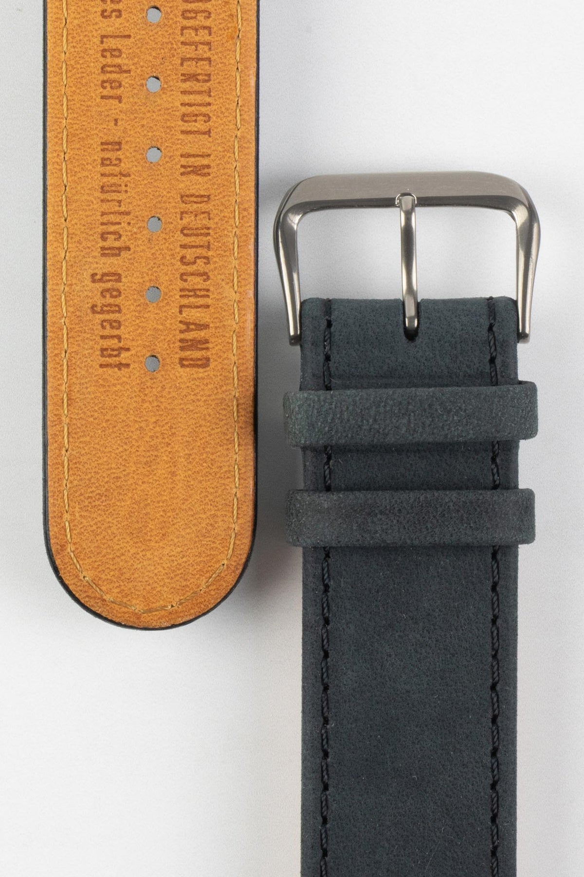 Close-up detail view of a dark navy nubuck leather watch strap, showcasing a soft suede-like texture, tonal stitching, dual keeper loops, and a polished stainless steel buckle, displayed alongside the natural tan leather lining embossed with “Handgefertigt in Deutschland” on a light grey background to highlight the premium handcrafted construction and contrasting underside.