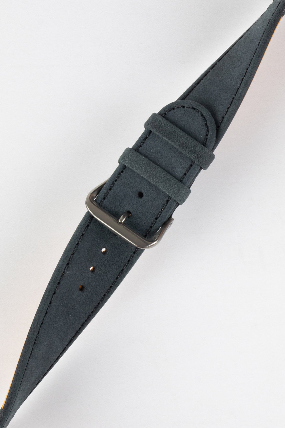 Close-up of a dark navy nubuck leather watch strap with a soft matte finish, tonal stitching, dual keeper loops, multiple adjustment holes, and a brushed stainless steel buckle, displayed diagonally on a light grey background to highlight the tapered profile, supple suede-like texture, and premium handcrafted construction.