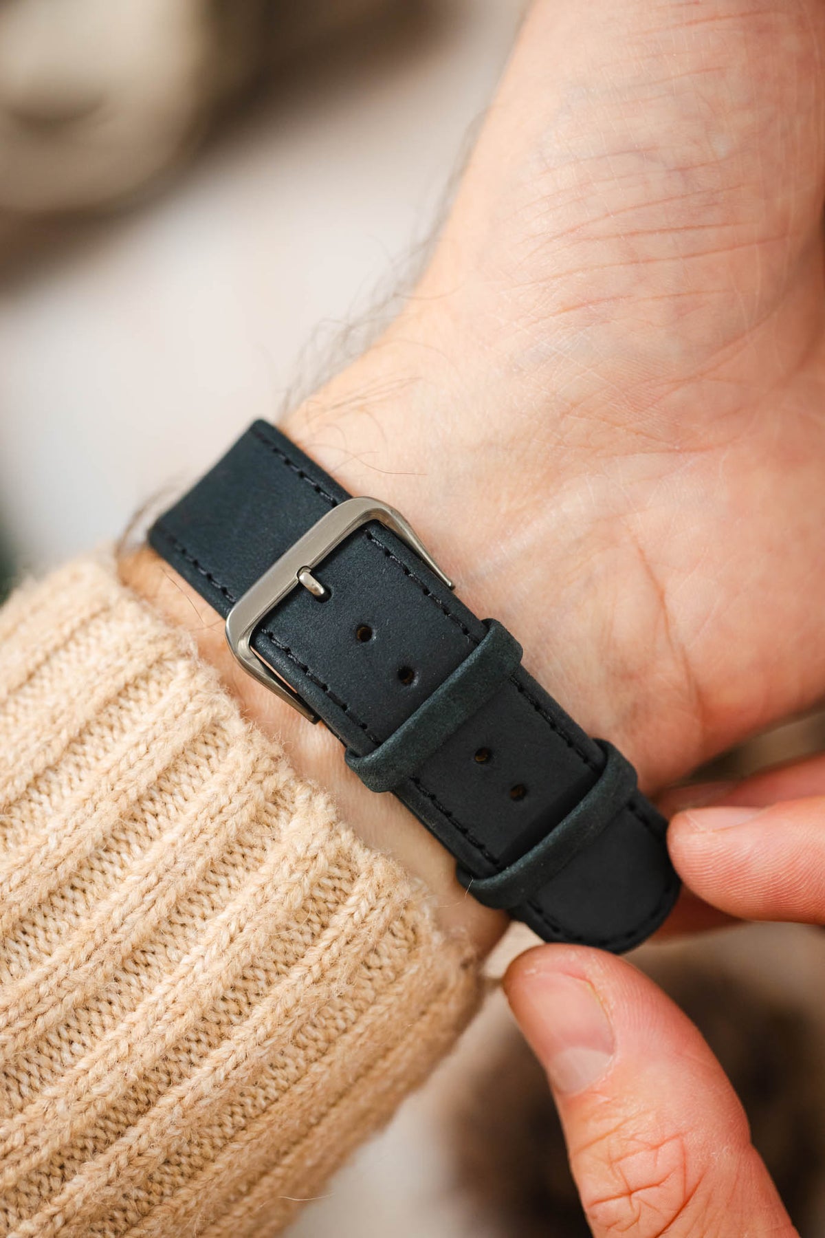 Close-up of a dark navy leather watch strap worn on the wrist, featuring a smooth matte finish, tonal black stitching, dual keeper loops, multiple adjustment holes, and a brushed stainless steel buckle, shown with a cream knitted sweater cuff to highlight the strap's minimalist design, supple leather construction, and comfortable everyday fit.