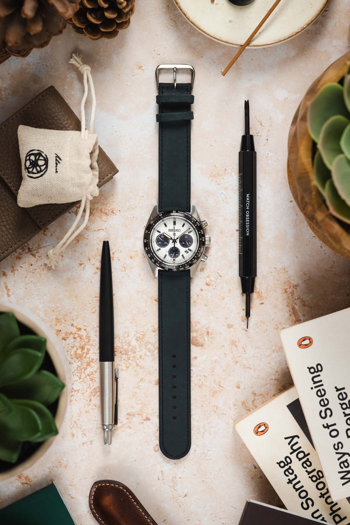 Seiko Speedtimer chronograph watch with a white panda dial, three black sub-dials, black tachymeter bezel, and a dark navy leather watch strap, displayed on a stone-effect surface alongside a Watch Obsession spring bar tool, pen, books, leather accessories, succulent plants, and decorative pine cones, highlighting the strap's smooth finish, minimalist design, and versatile everyday styling.