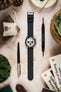 Seiko Speedtimer chronograph watch with a white panda dial, three black sub-dials, black tachymeter bezel, and a dark navy leather watch strap, displayed on a stone-effect surface alongside a Watch Obsession spring bar tool, pen, books, leather accessories, succulent plants, and decorative pine cones, highlighting the strap's smooth finish, minimalist design, and versatile everyday styling.